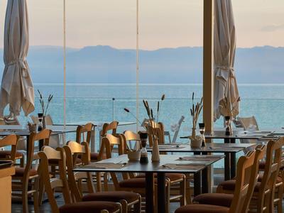 Blick durch ein Restaurant mit Holztischen und Stühlen auf das ruhige Meer bei Sonnenuntergang.