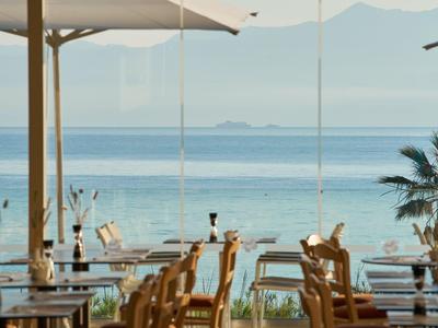 Restaurant mit Blick aufs Meer, draußen Tische, Stühle und Sonnenschirme bei klarem Himmel.