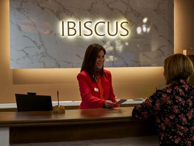 Rezeptionistin im roten Blazer bedient Gast am Hoteltheken-Schalter vor "IBISCUS" Schild.