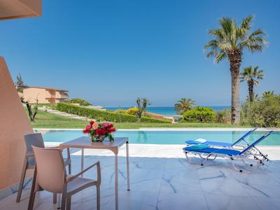 Sunny poolside with chairs, table, flowers, palm trees, and view of ocean under clear blue sky.