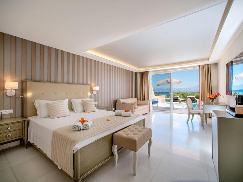 Bright hotel room with two beds, seating area, and glass doors opening to a balcony with sea view.