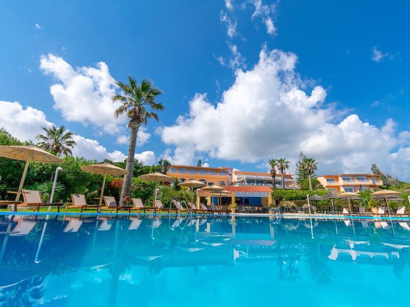 Bright blue pool in front of a hotel with palm trees under a partly cloudy sky.