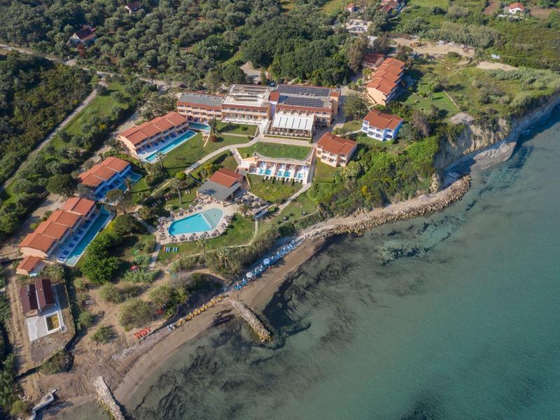 A coastal resort with multiple buildings, swimming pools, and green surroundings next to clear blue water.