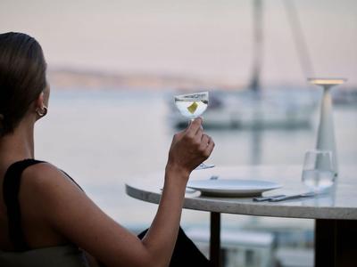 Mujer relajándose con copa de cóctel en mesa con vista al agua y barcos.