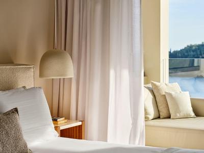 Heller Hotelzimmerbereich mit modernem Bett, Vorhängen und Sitzbereich mit Kissen neben Fenster.