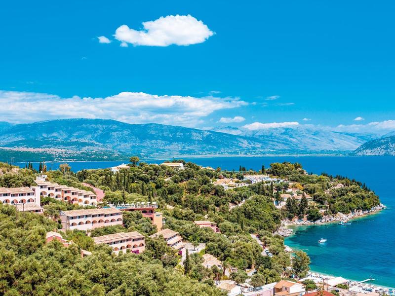 Küstenlandschaft mit grünen Hügeln, Häusern, blauem Meer und Bergen im Hintergrund unter klarem Himmel.