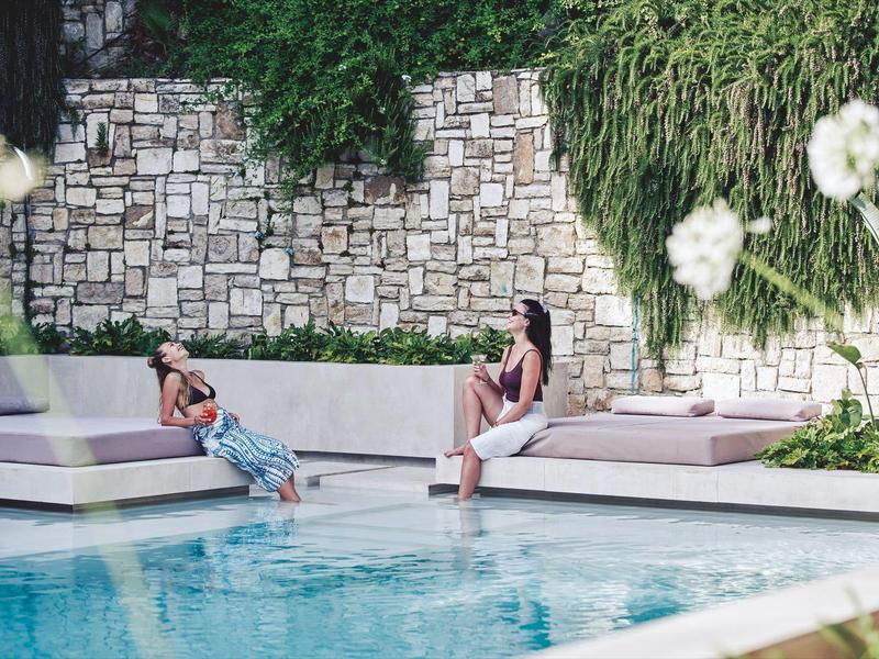 Zwei Frauen entspannen an einem blauen Pool mit weißen Liegen und Steinmauer im Hintergrund.