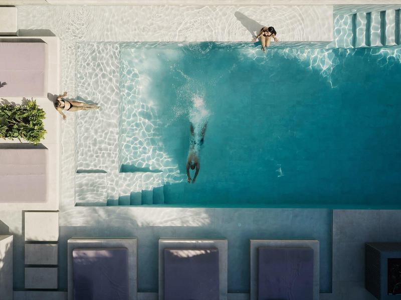 Vogelperspektive eines blauen Pools mit Springenden, Liegen und Pflanzen in rechteckiger Anordnung.