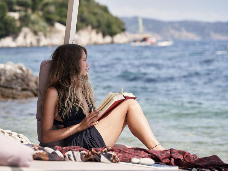 Frau sitzt entspannt am Strand, liest Buch, Meer und Felsen im Hintergrund.