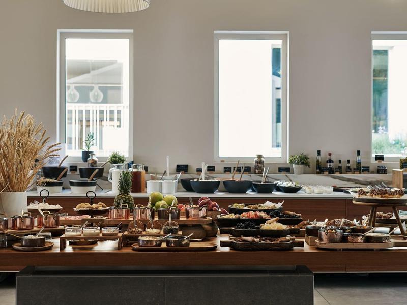 Buffet mit vielfältigem Essen auf Holztischen in hellem Raum mit drei großen Fenstern.