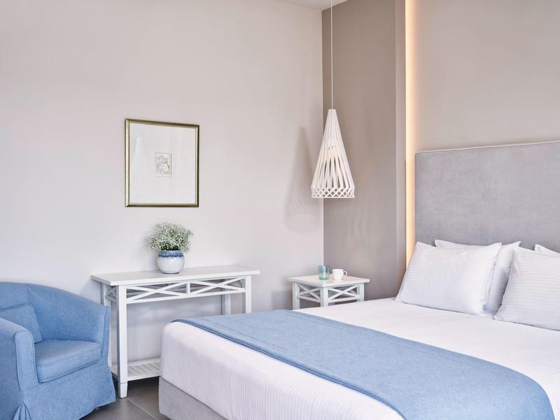 Helles Schlafzimmer mit grauem Bett, blauem Sessel, weißem Schreibtisch und zarter Wanddeko.