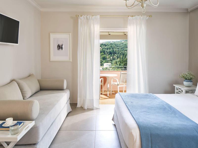 Helles Schlafzimmer mit Doppelbett, beigem Sofa, Fernseher und Terrasse mit Holztisch und Stühlen.