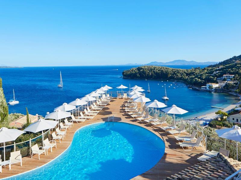 Pool mit blauer Wasserfläche, weißen Liegen und Sonnenschirmen am Meer mit Segelboot und Hügeln.
