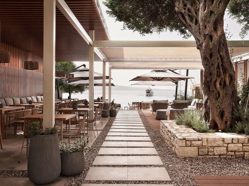 Moderner Außenbereich mit Sitzgelegenheiten, großen Bäumen und Wegplatten zu Sonnenschirmen am Wasser.