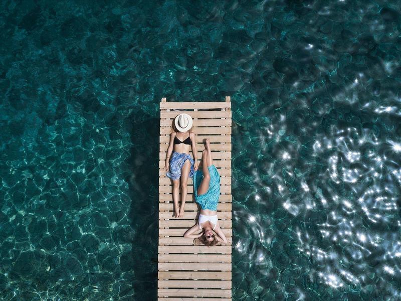 Zwei Personen liegen auf einem Steg über klarem, türkisfarbenem Wasser.