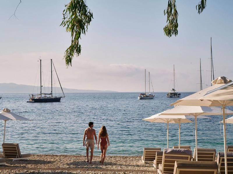 Zwei Personen gehen am Strand mit Segelbooten auf ruhigem Meer im Hintergrund.