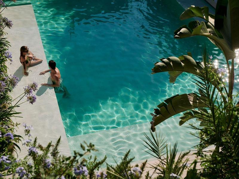 Zwei Personen entspannen im Wasser eines Pools, umgeben von grünen Pflanzen und Blumen.