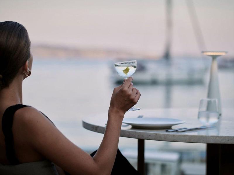Frau hält Cocktailglas und blickt auf ruhiges Wasser mit Segelboot.