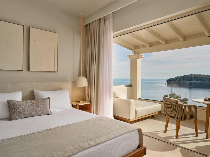 Helles Hotelzimmer mit großem Bett und Blick auf das Meer und eine Terrasse mit Sitzgelegenheiten.