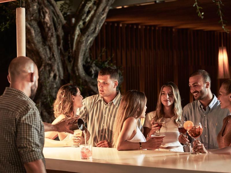 Sechs Personen unterhalten sich und trinken Getränke an einer beleuchteten Bar in gemütlicher Atmosphäre.