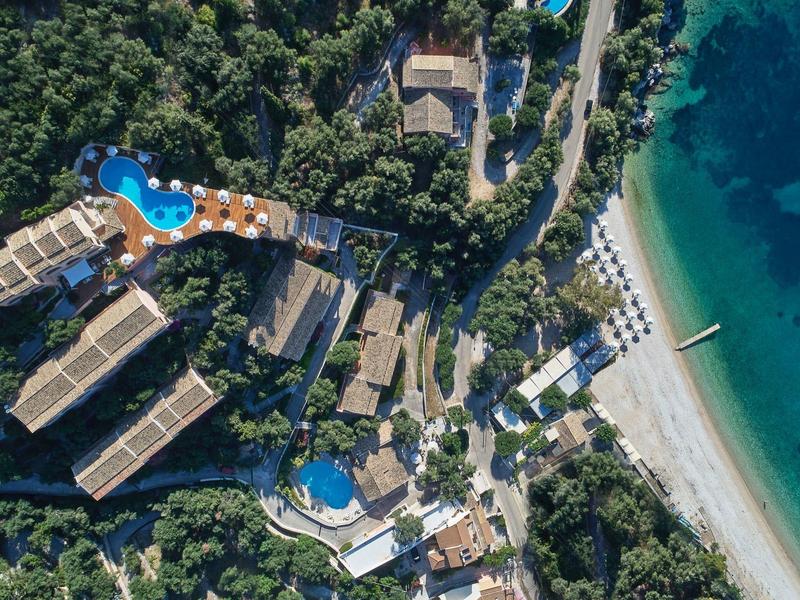 Luftaufnahme eines Hotels mit Pools, grünen Wäldern und sandigem Strand am türkisfarbenen Meer.