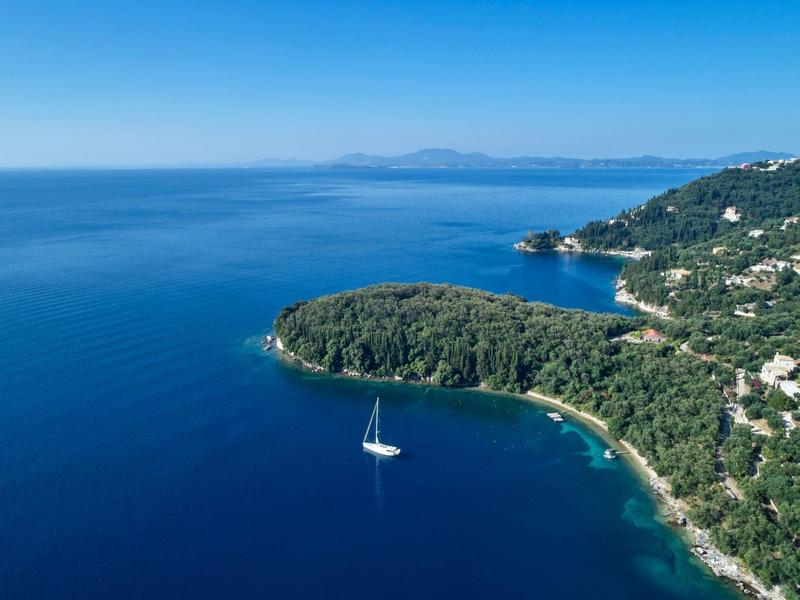 Küstenlinie mit grün bewaldeten Hügeln, weißen Häusern und Segelboot auf tiefblauem Meer.