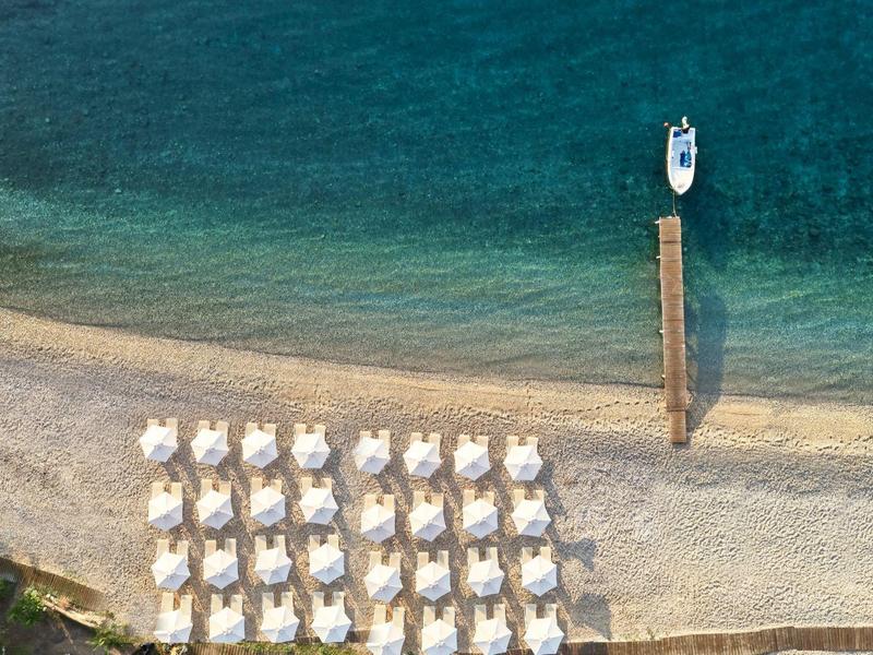 Luftaufnahme eines Strandes mit Reihen von weißen Sonnenschirmen und einem Steg mit Boot im klaren Wasser.