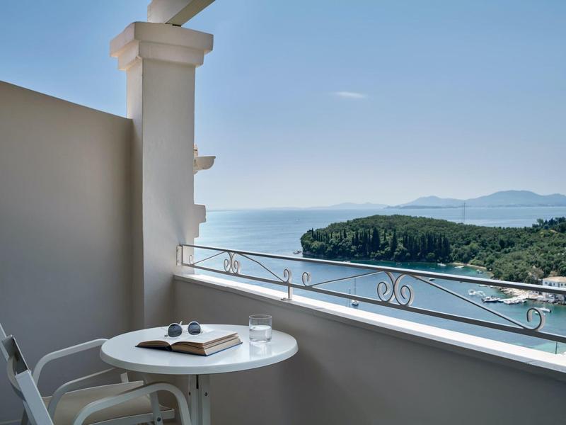 Balkon mit kleinem weißem Tisch, zwei Stühlen und Meerblick auf grüne Inseln und blauen Himmel.
