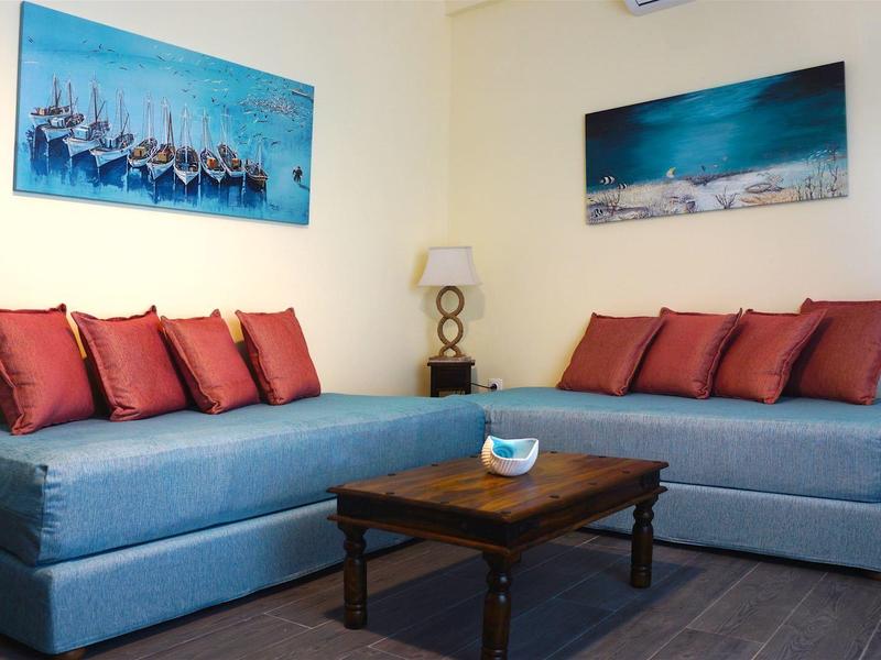 Wohnzimmer mit blauen Sofas, roten Kissen, Holztisch und maritimen Bildern an weißen Wänden.