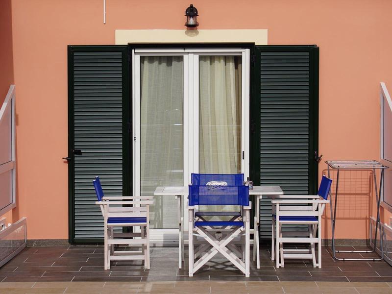 Vier weiße Stühle mit blauen Sitzen rund um blauen Tisch vor Haus mit grünem Fensterladen.
