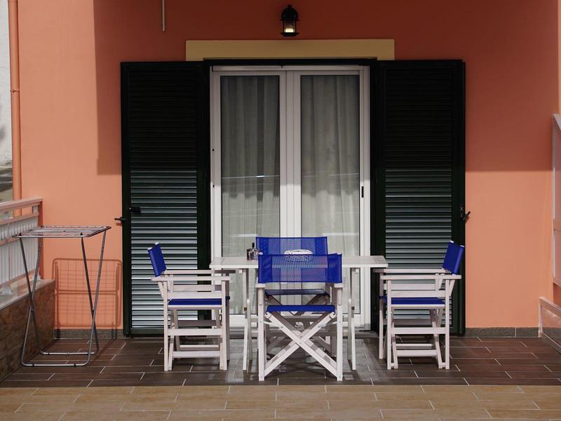 Terrasse mit weißem Gartentisch, blauen Stühlen vor einem Fenster mit schwarzen Rollläden.