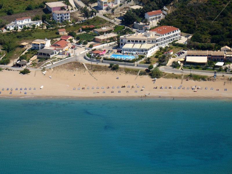 Flacher Sandstrand mit Sonnenschirmen, dahinter Gebäude und grüne Vegetation am Meer.