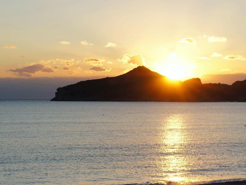 Sonnenuntergang über dem Meer mit Hügeln im Hintergrund und ruhigem Wasser.