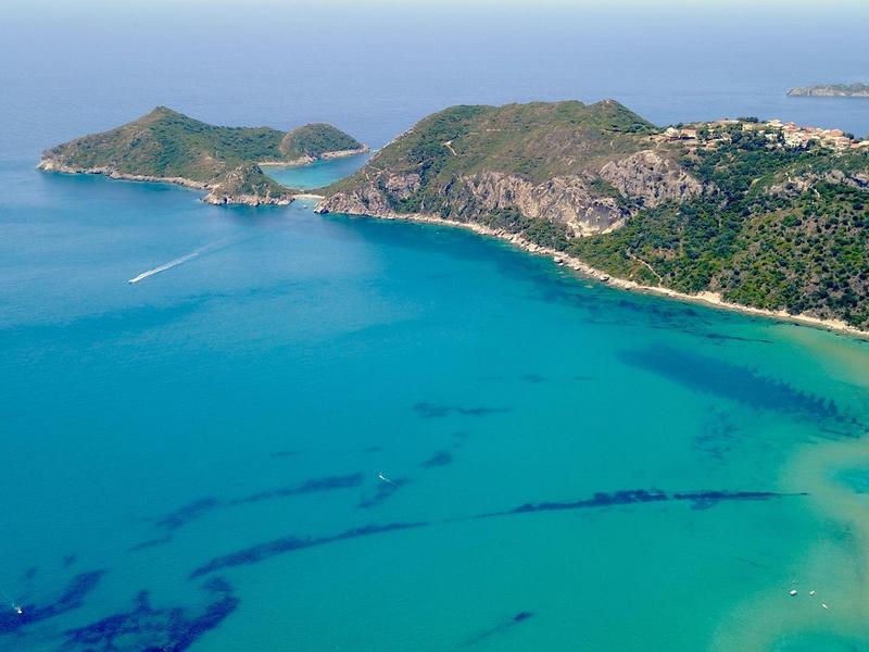 Luftaufnahme einer Türkis-blaue Bucht mit sauberem Sandstrand und bewaldeten Hügeln.