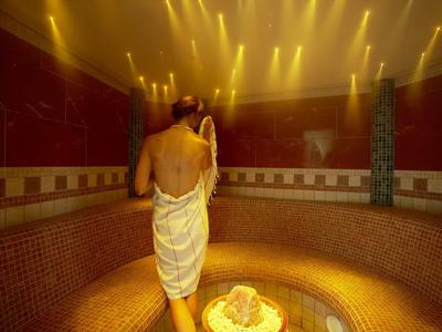 Ein Mann in einem weißen Handtuch betritt einen dampfigen Spa-Raum mit warmem Licht und Mosaikfliesen.