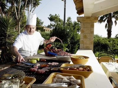 Koch in weißer Kleidung grillt verschiedene Fleischsorten draußen auf großem Grill unter Sonne.