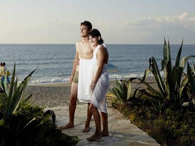 Ein Paar am Strand, bedeckt mit weißen Handtüchern, umgeben von Pflanzen und Meer im Hintergrund.