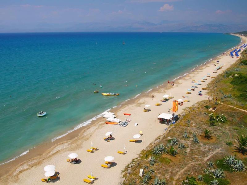 Langer Sandstrand mit Sonnenschirmen, Liegestühlen, grünem Wasser und bewachsenem Küstenhügel.