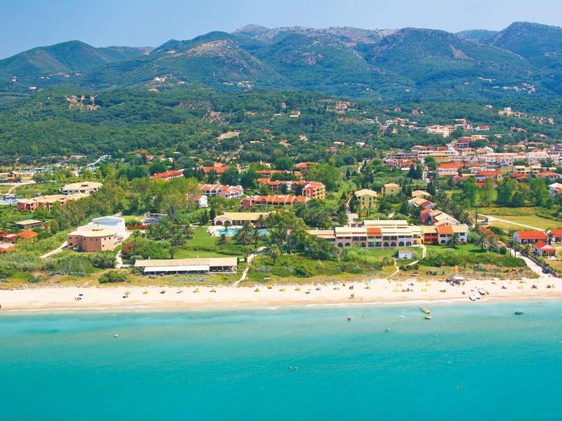 Blick auf einen sonnigen Sandstrand mit blauem Meer, dahinter grüne Vegetation und Berge im Hintergrund.
