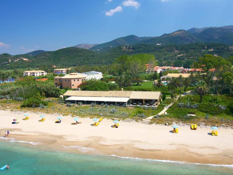 Strand mit Sand, Sonnenschirmen, türkisfarbenem Meer und grünen Hügeln im Hintergrund.