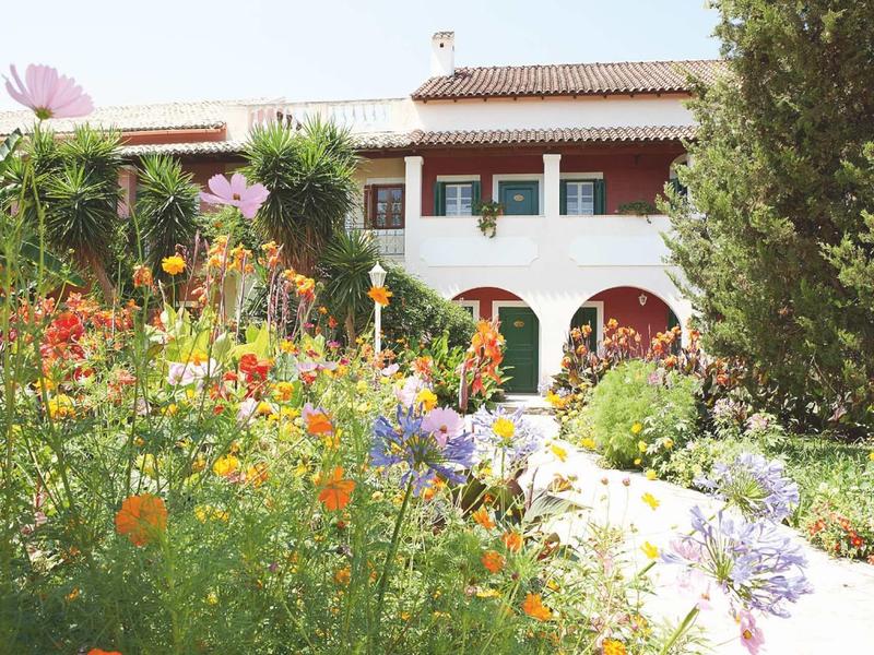 Haus mit weißer Fassade und rotem Dach, umgeben von bunten Blumen und grünen Büschen.