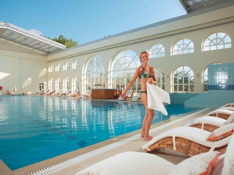 Helle Schwimmhalle mit großem Pool, Liegestühlen und einer Frau mit weißem Handtuch.