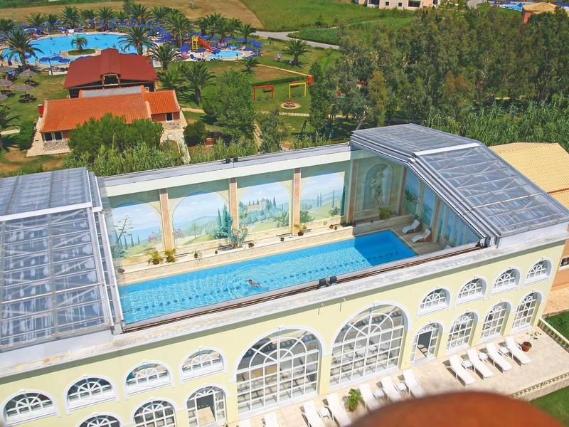 Großes Gebäude mit Glasdach und langem Pool, umgeben von grünem Gelände und Gebäuden im Hintergrund.