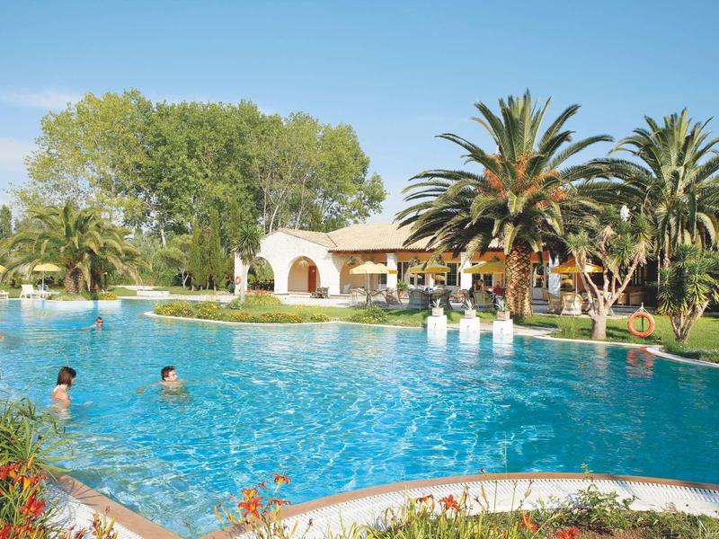 Großer blauer Pool mit Menschen, umgeben von Palmen und einem hellen Gebäude im sonnigen Garten.