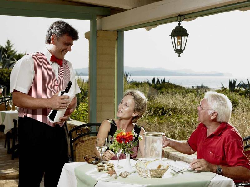 Ein Kellner serviert zwei älteren Personen an einem gedeckten Tisch auf einer Terrasse mit Meerblick.