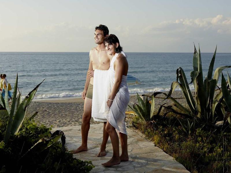 Ein Paar am Strand, bedeckt mit weißen Handtüchern, umgeben von Pflanzen und Meer im Hintergrund.