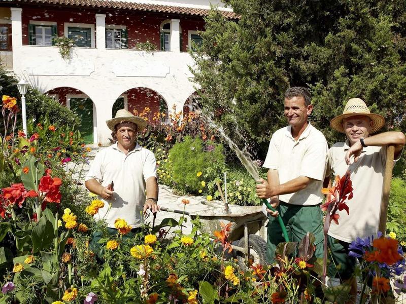 Drei Personen gießen bunte Blumen in einem sonnigen Garten vor einem Haus mit Balkon.