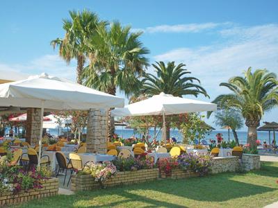 Außenbereich eines Hotels mit Tischen unter Sonnenschirmen, umgeben von Palmen und Blick aufs Meer.