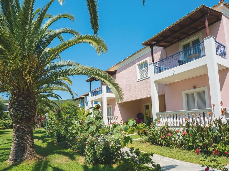 Rosa Ferienanlage mit Balkonen und Palmen in gepflegtem Garten unter blauem Himmel.