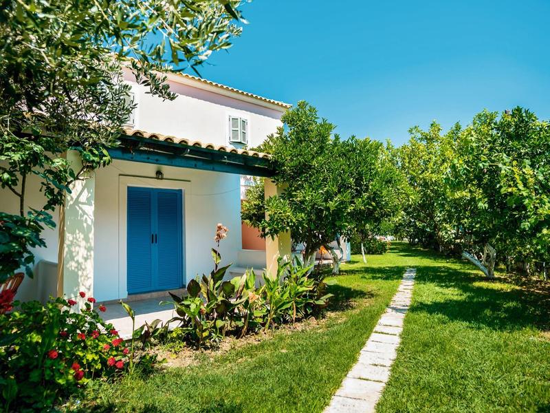 Weißes Haus mit blauen Türen in einem Garten mit grünen Bäumen und Steinweg.